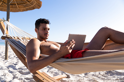 Caucasian man enjoying time at the beach, lying on a hammock and using a digital tablet