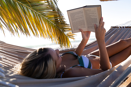 Caucasian woman enjoying time at the beach, lying on a hammock and reading a book