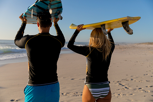 Caucasian couple enjoying time at the beach, holding surfboards above their heads, walking towards the sea