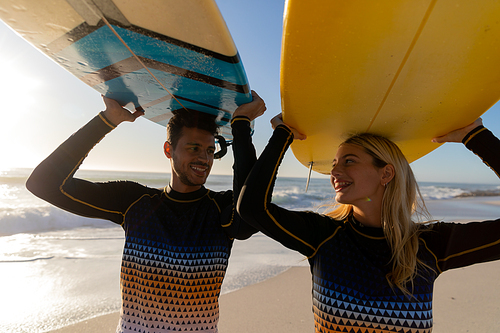 Caucasian couple enjoying time at the beach on a sunny day, holding surfboards above their heads with sea in the background