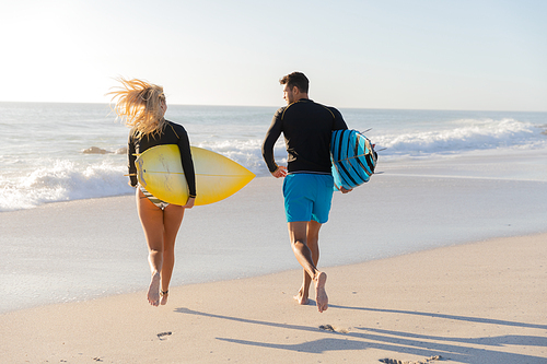 Caucasian couple enjoying time at the beach on a sunny day, holding surfboards and walking with sea in the background