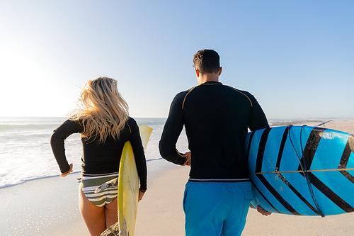 Caucasian couple enjoying time at the beach on a sunny day, holding surfboards and walking with sea in the background