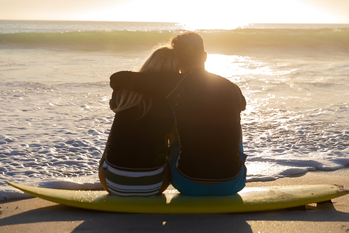 Caucasian couple enjoying time at the beach during a pretty sunset, sitting on a surfboard and embracing with sea in the background