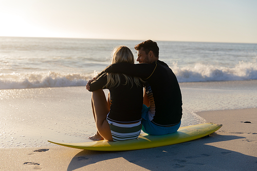 Caucasian couple enjoying time at the beach during a pretty sunset, sitting on a surfboard and embracing with sea in the background