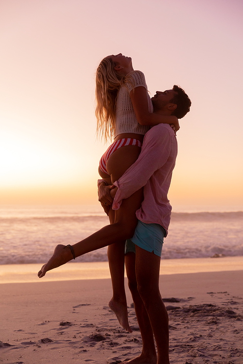 Caucasian couple enjoying time at the beach during a pretty sunset, a man is lifting a woman with blue sky and sea in the background