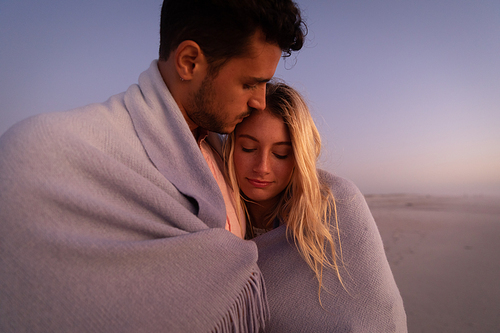 Caucasian couple enjoying time at the beach, embracing and covering themselves with a blanket, a man is kissing a woman on her forehead
