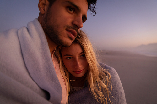 Caucasian couple enjoying time at the beach during a pretty sunset, embracing and covering themselves with a blanket, with yellow sand in the background