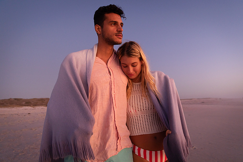 Caucasian couple enjoying time at the beach during a pretty sunset, embracing and covering themselves with a blanket, with yellow sand in the background