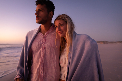 Caucasian couple enjoying time at the beach during a pretty sunset, embracing and covering themselves with a blanket, with yellow sand in the background