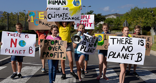 Front view of a diverse group of elementary school pupils walking down a road in the sun on a protest march, carrying signs with environmental and conservation slogans on them, one girl shouting in a megaphone