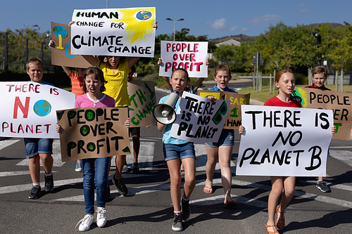 Front view of a diverse group of elementary school pupils walking down a road in the sun on a protest march, carrying signs with environmental and conservation slogans on them, one girl holding a megaphone