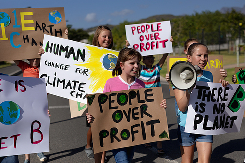 Front view of a diverse group of elementary school pupils walking down a road in the sun on a protest march, carrying signs with environmental and conservation slogans on them, one girl holding a megaphone