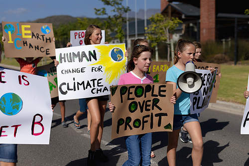 Front view of a diverse group of elementary school pupils walking down a road in the sun on a protest march, carrying signs with environmental and conservation slogans on them, one girl shouting in a megaphone