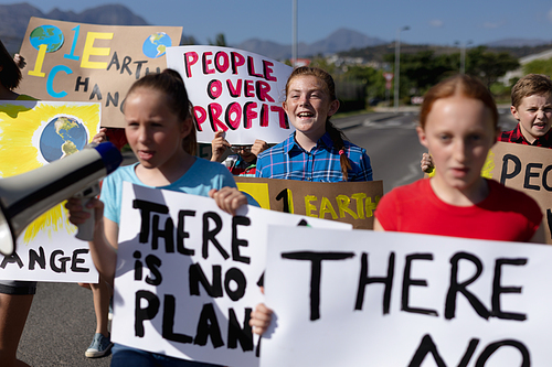 Front view of a diverse group of elementary school pupils walking down a road in the sun on a protest march, carrying signs with environmental and conservation slogans on them, and one girl holding a megaphone