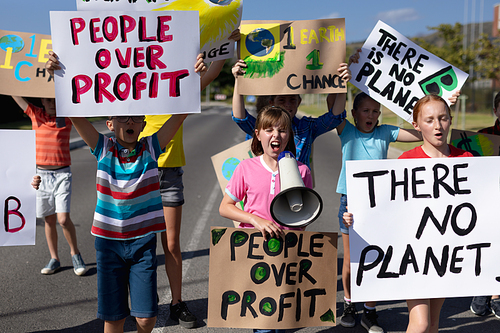 Front view of a diverse group of elementary school pupils on a protest march, carrying signs with environmental and conservation slogans on them, and one girl shouting in a megaphone as they walk down a road in the sun