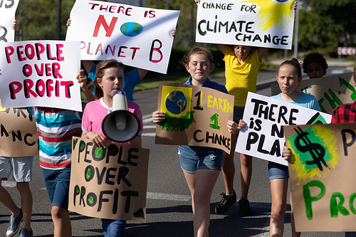 Front view of a diverse group of elementary school pupils on a protest march, carrying signs with environmental and conservation slogans on them, one girl shouting in a megaphone as they walk down a road in the sun