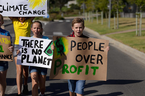 Front view of Caucasian male and female elementary school pupils on a protest march, carrying signs with environmental and conservation slogans on them, shouting and walking down a road in the sun