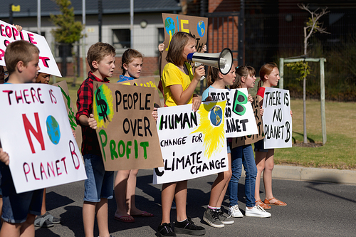 Side view of a group of Caucasian elementary school pupils on a protest march, carrying signs with environmental and conservation slogans on them standing in a road in the sun, one girl shouting in a megaphone