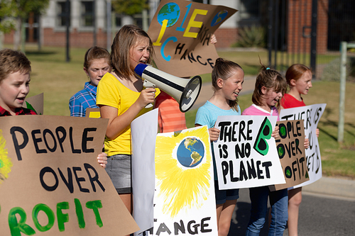 Side view of a group of Caucasian elementary school pupils on a protest march, carrying signs with environmental and conservation slogans on them, and one girl shouting in a megaphone as they walk down a road in the sun