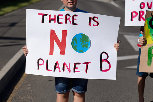 Front view mid section of a Caucasian elementary school boy on a protest march, carrying a sign with environmental conservation slogan on it, with his classmates carrying signs beside him walking down a road in the sun