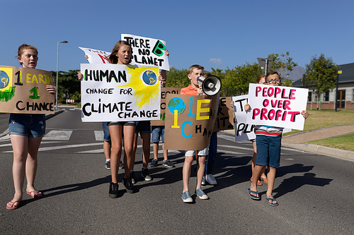 Front view of a group of Caucasian elementary school pupils on a protest march, carrying signs with environmental and conservation slogans on them, and one boy shouting in a megaphone while they walk down a road in the sun
