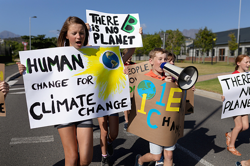 Front view close up of a group of Caucasian elementary school pupils on a protest march, carrying signs with environmental and conservation slogans on them, and one boy shouting in a megaphone while they walk down a road in the sun