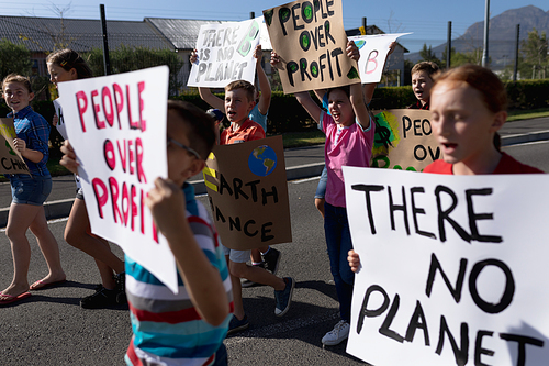 Side view of a diverse group of elementary school pupils on a protest march, carrying signs with environmental and conservation slogans on them and shouting, walking down a road in the sun