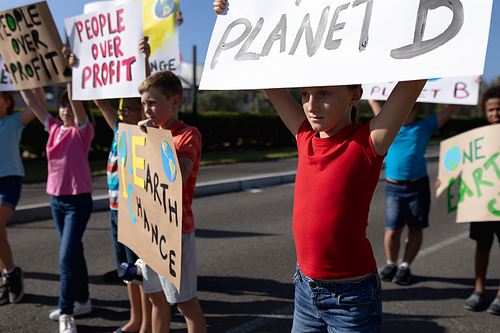 Front view of a diverse group of elementary school pupils on a protest march, carrying signs with environmental and conservation slogans on them and walking down a road in the sun