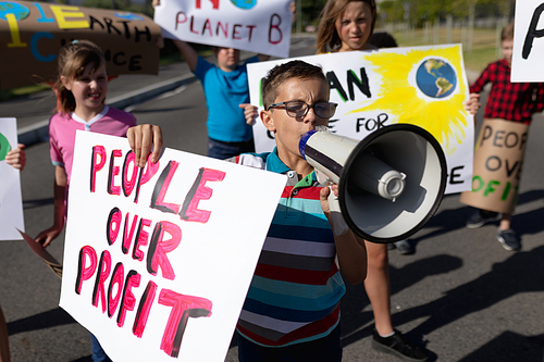 Front view of a group of Caucasain elementary school pupils on a protest march, carrying signs with environmental and conservation slogans on them, and a boy wearing glasses in the foreground shouting in a megaphone