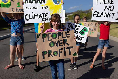 Front view of a group of Caucasian elementary school pupils walking down a road in the sun on a protest march, carrying signs with environmental and conservation slogans on them
