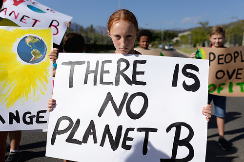 Portrait of a Caucasian girl carrying a sign with a slogan on it and walking down a road in the sun with a diverse group of elementary school pupils also carying signs on an environmental and conservation protest march