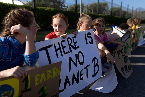 Side view of a group of Caucasian elementary school pupils on a protest march, carrying signs with environmental and conservation slogans on them, walking down a road in the sun