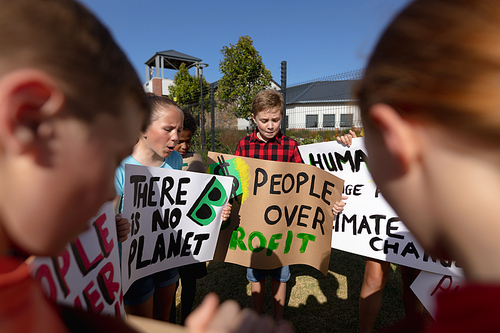 Over the shoulder view of a group of Caucasian elementary school pupils on a protest march, carrying signs with environmental and conservation slogans on them, walking down a road in the sun