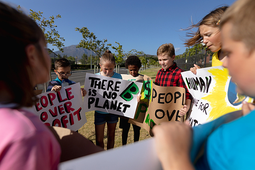 Over the shoulder view of a diverse group of elementary school pupils on a protest march, carrying signs with environmental and conservation slogans on them, walking down a road in the sun