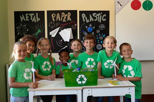 Portrait of a diverse group of eight schoolchildren wearing green t shirts with a white recycling logo on them standing around a recycling box on a table and holding three miniature wind turbines in front of posters about recyclable materials and smiling to camera in an elementary school classroom