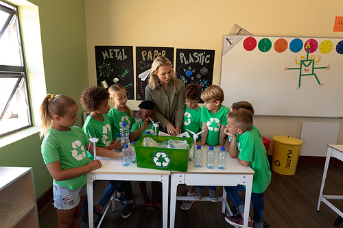 Front view of a Caucasian female school teacher with long blonde hair and a diverse group of schoolchildren wearing green t shirts with a white recycling logo on them standing around a recycling box on a table, holding minature wind turbines and discussing recyclable materials during a lesson in an elementary school classroom