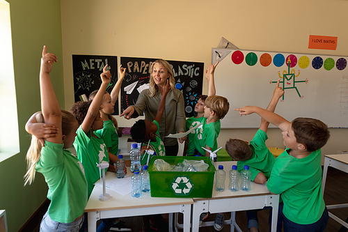 Front view of a Caucasian female school teacher with long blonde hair and a diverse group of schoolchildren wearing green t shirts with a white recycling logo on them standing around a recycling box, miniature wind turbines and recyclable materials on a table during a lesson in an elementary school classroom, the children raising their hands to answer a question and the teacher pointing