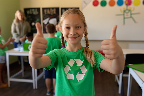 Portrait of a Caucasian schoolgirl wearing a green t shirt with a white recycling logo on it, looking to camera, smiling with her thumbs up in an elementary school classroom, with her classmates and teacher standing in the background
