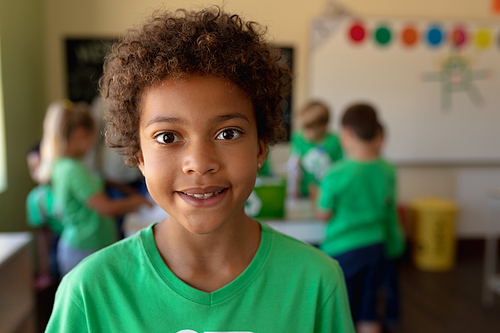 Portrait close up of an African American schoolgirl with short hair wearing a green t shirt and smiling to camera in an elementary school classroom, with her  classmates and teacher standing in the background