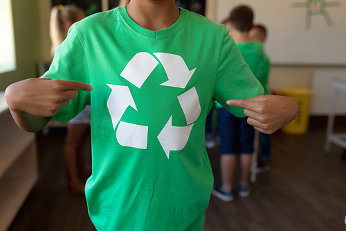 Front view mid section of an African American schoolgirl wearing a green t shirt with a white recycling logo on it and pointing at it, in an elementary school classroom, with her classmates standing in the background