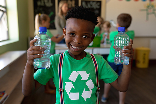 Portrait of an African American schoolboy with short hair wearing a green t shirt with a white recycling logo on it, holding two plastic water bottles and looking to camera, smiling, in an elementary school classroom, with his classmates and teacher standing in the background
