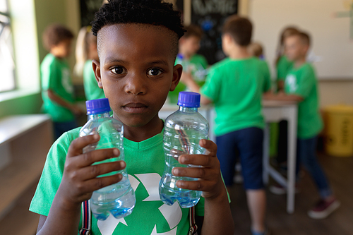 Portrait close up of an African American schoolboy with short hair wearing a green t shirt with a white recycling logo on it, holding two plastic water bottles and looking to camera in an elementary school classroom, with his classmates standing in the background