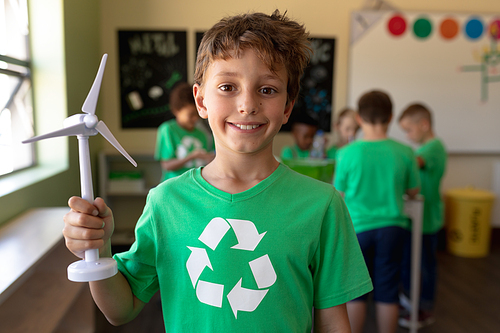 Portrait close up of a Caucasian schoolboy with short dark hair wearing a green t shirt with a white recycling logo on it, holding miniature wind turbine and looking to camera smiling in an elementary school classroom, with his classmates standing in the background