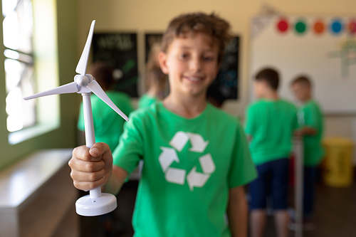 Front view of a Caucasian schoolboy with short dark hair wearing a green t shirt with a white recycling logo on it, holding miniature wind turbine and looking to camera smiling in an elementary school classroom, with his classmates standing in the background, focus on the foreground
