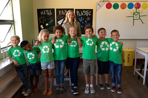 Portrait of a Caucasian female school teacher with long blonde hair and a diverse group of eight schoolchildren wearing green t shirts with a white recycling logo on them, standing in an elementary school classroom with arms around each other, smiling to camera