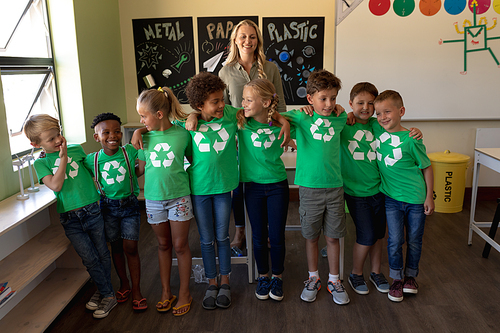 Front view of a Caucasian female school teacher with long blonde hair and a diverse group of eight schoolchildren wearing green t shirts with a white recycling logo on them standing in an elementary school classroom with arms around each other, looking at each other and smiling