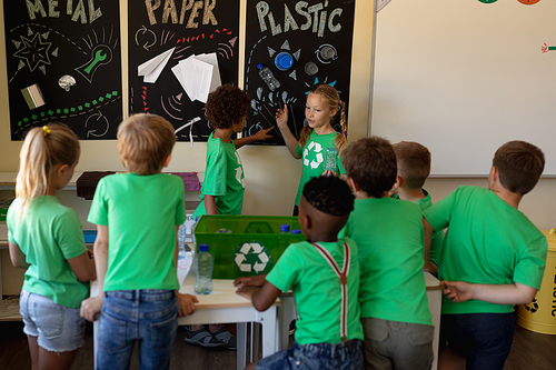 Rear view of a diverse group of schoolchildren wearing green t shirts standing around a recycling box and listening to a Caucasian and an African American schoolgirl standing and talking to them and pointing to a poster about recyclable materials in an elementary school classroom