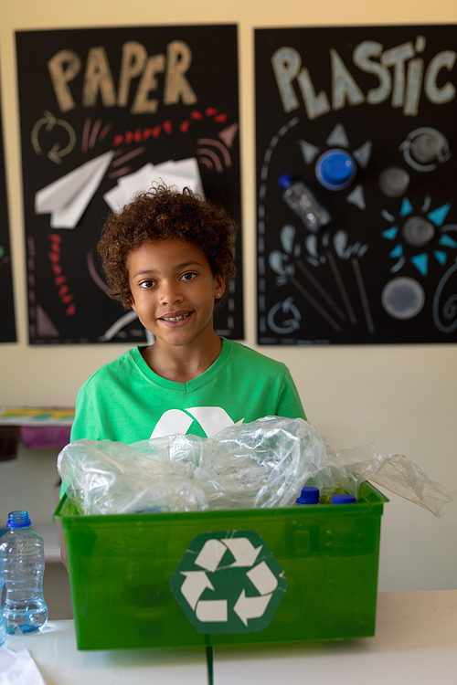 Portrait of an African American schoolgirl with short hair wearing a green t shirt with a white recycling logo on it, holding a recycling crate and looking to camera smiling in an elementary school classroom, with poster about recyclable materials in the background