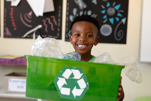 Portrait of an African American schoolboy with short hair holding a green crate with a white recycling logo on it and looking to camera smiling in an elementary school classroom