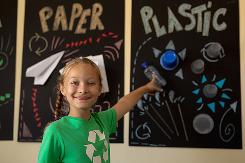 Portrait of a Caucasian schoolgirl wearing a green t shirt with a white recycling logo on it, pointing to a poster about recyclable materials and looking to camera smiling in an elementary school classroom